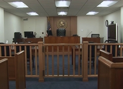 [ai] An empty courtroom featuring wooden benches, a judge's chair at the front, and various desks for court personnel. The room is well-lit with a seal on the wall and an American flag in the background.